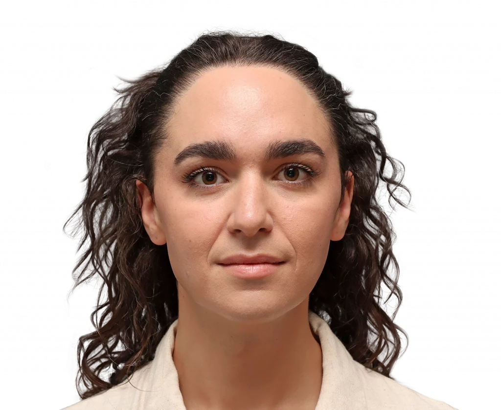 Close-up portrait of a woman facing the camera with brown eyes and wavy dark hair, wearing a light beige shirt against a black background.